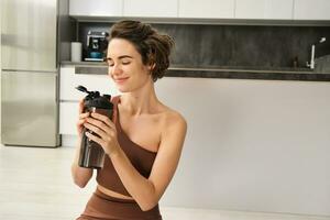 aptitude instructeur, souriant Jeune femme faire des exercices à maison, est assis sur yoga tapis et les boissons eau, Est-ce que des exercices, formation session dans sa cuisine photo