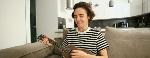 portrait de Jeune moderne femme, musicien en jouant ukulélé à maison, cueillette accords pour Nouveau chanson, séance sur canapé dans vivant pièce et souriant photo