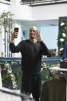 achats centre commercial boutique arabe homme client agitant salut tandis que fabrication selfie sur téléphone intelligent. Jeune souriant blogueur salutation social médias suiveurs tandis que vivre diffusion dans Vêtements boutique photo