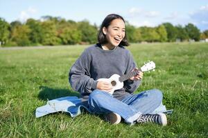 content brunette asiatique fille, étudiant en jouant ukulélé séance sur herbe dans parc, relaxant, en chantant chanson, mode de vie concept photo