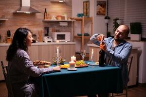 de bonne humeur mari ouverture une bouteille de du vin pendant romantique dîner avec épouse dans cuisine. content couple en parlant, séance à table dans cuisine, profiter le repas, célébrer leur anniversaire . photo