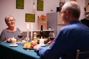 mature femme ayant une conversation avec mari tandis que en portant verre de rouge du vin dans cuisine. Sénior couple séance à le table dans cuisine, en parlant, profiter le repas, célébrer leur anniversaire dans le à manger chambre. photo