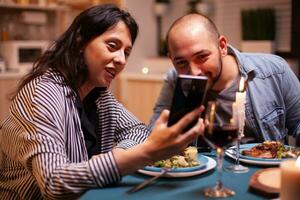 content couple en train de regarder vidéo sur téléphone intelligent dans cuisine pendant anniversaire. adultes séance à le table dans le cuisine navigation, recherche, en utilisant smartphone, l'Internet, célébrer anniversaire. photo