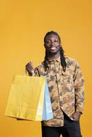 souriant élégant homme en portant achats papier Sacs après fabrication achat sur remise vente à vente au détail magasin. africain américain Jeune adulte porter paquets après achat élégant des choses dans studio photo