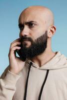 sérieux arabe homme parlant sur téléphone intelligent et à la recherche loin. Jeune chauve barbu arabe la personne répondre mobile téléphone appel et écoute à interlocuteur avec tendu faciale expression photo