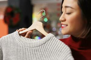 acheteur en cours d'analyse vêtements sur cintres avant achat cadeaux pour Noël veille fête, femme à la recherche à chandail tissu. asiatique client vérification Nouveau Ventes marchandise dans magasin. proche en haut. photo