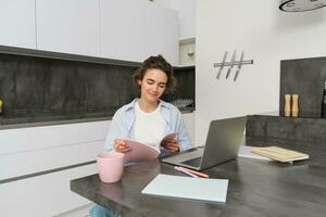 portrait de Jeune magnifique femme, études à maison, flips sa cahier d'exercices, travaux de maison, les usages portable à apprendre sur télécommande, fait du Remarques pendant séminaire en ligne photo