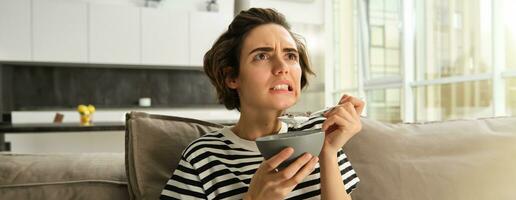 image de femme en mangeant petit déjeuner dans de face de la télé, à la recherche à écran avec inquiet affronter, en portant bol de céréales avec Lait et une cuillère, dépenses temps dans vivant pièce photo