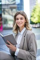 une Jeune femme dans entreprise costume, est assis avec carnet et stylo, prend Remarques, travaux et écrit vers le bas sa des idées photo