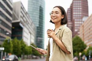 content femme explorant le ville. Jeune coréen fille détient tablette, les boissons café et des promenades le long de rue avec gros sourire sur sa visage photo
