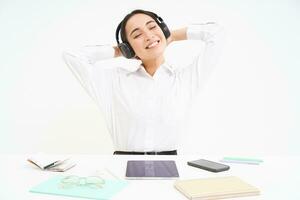 souriant femme d'affaires dans écouteurs, est assis à Bureau bureau écoute la musique et se détend, repose après travail, blanc Contexte photo