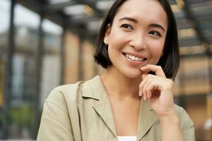 proche en haut portrait de asiatique fille avec content affronter, souriant et en riant, émouvant sa peau, permanent sur rue sur ensoleillé journée photo