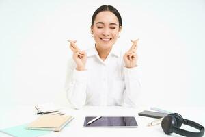 optimiste asiatique femme d'affaires, traverser les doigts pour bien chance, est assis dans lieu de travail Bureau et fait du souhait, blanc Contexte photo