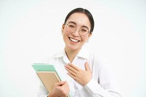 amical content asiatique femme dans lunettes, prof met main sur poitrine et sourires, présente se, des stands plus de blanc Contexte photo