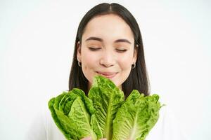 proche en haut portrait de content coréen femme, spectacles sa visage avec laitue, en mangeant chou, aime légumes, se sent en bonne santé et sous tension, blanc Contexte photo