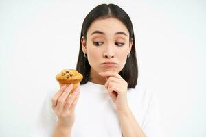 image de pensif coréen femme, regards à petit gâteau avec réfléchi affronter, en pensant à propos dessert, blanc Contexte photo