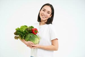 souriant asiatique femme, montrant sa biologique vert légumes, en mangeant en bonne santé brut nourriture, blanc Contexte photo