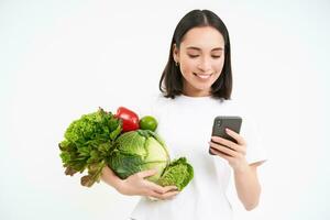 portrait de coréen femme détient légumes, ordres vert biologique nourriture sur téléphone intelligent application, blanc Contexte photo