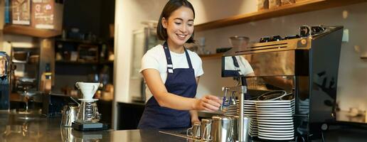 souriant fille barista dans café, en train de préparer cappuccino dans café machine, fumant lait, portant uniforme tablier photo