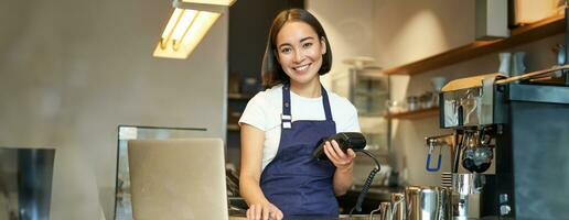 souriant asiatique barman, fille avec carte Terminal, Paiement machine et ordinateur portable, permanent dans café, En traitement Paiement pour café commande photo