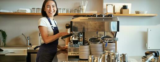 content souriant barman, barista en utilisant café machine, préparer commande, fabrication cappuccino ou latté, portant bleu tablier photo