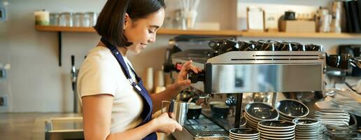 souriant Jeune asiatique barista fille, en utilisant café machine bateau à vapeur, fumant Lait pour cappuccino commande, permanent dans tablier, travail dans café photo