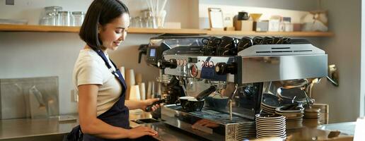 content magnifique asiatique fille, barista dans tablier travaux avec café machine derrière comptoir, permanent dans café et fabrication cappuccino photo