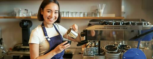 souriant asiatique barista fille, fabrication café, verser à la vapeur Lait dans cappuccino, Faire latté art dans tasse, travail dans café photo