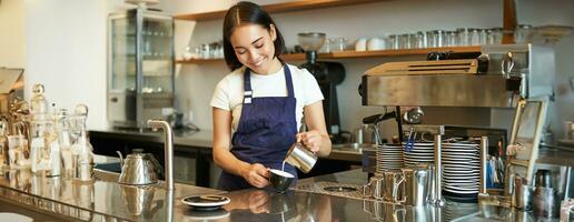 mignonne coréen barista fille, verser Lait dans café, préparer cappuccino avec latté art, travail dans café derrière comptoir. gens et lieu de travail photo