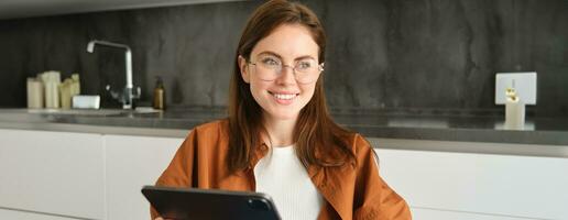 portrait de Jeune magnifique femme dans lunettes, travail de maison, en train d'étudier en ligne dans sa cuisine, en portant numérique tablette, en ligne achats photo