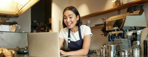 enthousiaste asiatique barman, fille dans tablier, à la recherche à ordinateur portable, vidéo chats, parlant en ligne, permanent derrière compteur dans café magasin photo