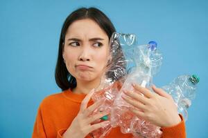 agacé coréen femme à la recherche avec blâmer affronter, en portant Plastique bouteilles, permanent plus de bleu studio Contexte photo