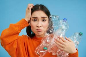 triste asiatique femme en portant Plastique bouteilles, à la recherche troublé et bouleversé, tri déchets pour recyclage, permanent plus de bleu Contexte photo