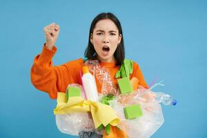 en colère coréen femme des cris, détient Plastique déchets, exhorte à recycler ordures, secoue poing, bleu Contexte photo