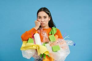 agacé Jeune femme nettoyage loger, en portant Plastique des ordures pour recyclage, fatigué de collecte déchets, permanent plus de bleu Contexte photo