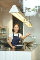 content asiatique fille barista travaux dans café boutique, vagues à client, En traitement ordres derrière compteur dans café, porte bleu tablier photo