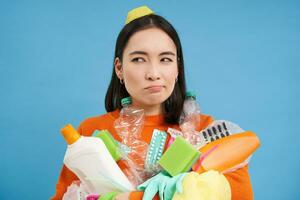 perplexe Jeune femme avec pile de ordures, détient Plastique bouteilles et poubelle pour recyclage, regards perplexe, bleu Contexte photo