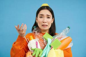 frustré asiatique femme pleurs, en portant charges de vide Plastique bouteilles et des ordures pour recyclage, à la recherche stressé, bleu Contexte photo