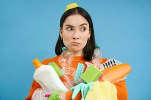 perplexe Jeune femme avec pile de ordures, détient Plastique bouteilles et poubelle pour recyclage, regards perplexe, bleu Contexte photo