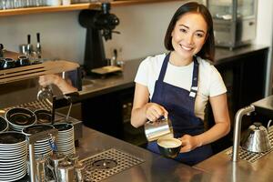 souriant asiatique fille barman, café Personnel verser à la vapeur Lait dans café, préparer cappuccino avec latté art, permanent dans bleu tablier derrière compteur photo