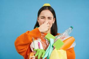 portrait de femme se ferme sa nez de dégoût, en portant puant ordures, sale Plastique articles et vide bouteilles avec mal odeur, permanent plus de bleu Contexte photo