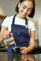 verticale coup de brunette asiatique fille barman, portant tablier, fabrication café, préparer commande dans café, verser Lait dans tasse pour cappuccino latté art photo