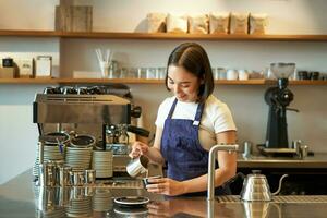 content souriant café propriétaire, fille barista dans tablier, fabrication cappuccino, latté art avec à la vapeur lait, permanent derrière compteur photo
