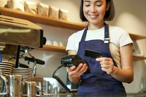 magnifique souriant asiatique fille, barista insérer client crédit carte dans pos Terminal, En traitement paiement, prise ordres dans café magasin photo