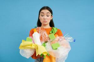 asiatique fille regards à sa mains en portant Plastique déchets, recyclage matériaux, collecte litière à recycler, bleu Contexte photo