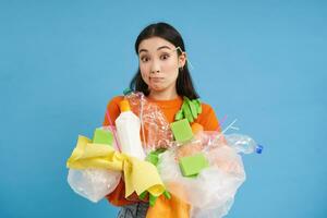 mignonne asiatique femme en portant Plastique déchets, des ordures pour recyclage, à la recherche Aucune idée et haussant les épaules, bleu Contexte photo