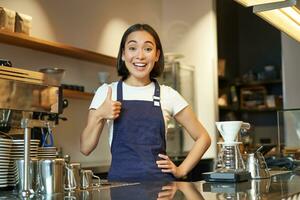 portrait de souriant coréen barman, fille à le comptoir, porte bleu tablier, travaux dans café boutique, spectacles les pouces en haut. gens à travail photo