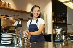 souriant fille barista dans café, en train de préparer cappuccino dans café machine, fumant lait, portant uniforme tablier photo