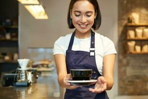 souriant coréen fille dans uniforme, travail dans café, donnant vous tasse de café, en train de préparer commande pour client photo