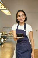 souriant magnifique asiatique fille barman, travail dans café, en portant tasse de thé, donnant client leur commande, portant uniforme tablier photo